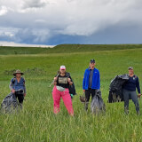 Volunteers stand in a green field with tools and large bags of weeds on shoulders