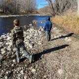 Two boys play by a river. The water is low and the trees have no leaves. It looks wintery but sunny.