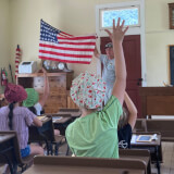 A student is raising their hand to answer questions in the schoolhouse as a volunteer holds a flag