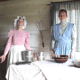 A mom and daughter volunteer team in 1880s' dresses and bonnets prepare food in a log house